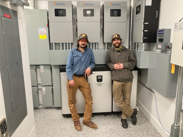 Two technicians posing in an electrical room with three Sol-Ark solar inverter cabinets on the wall behind them.
