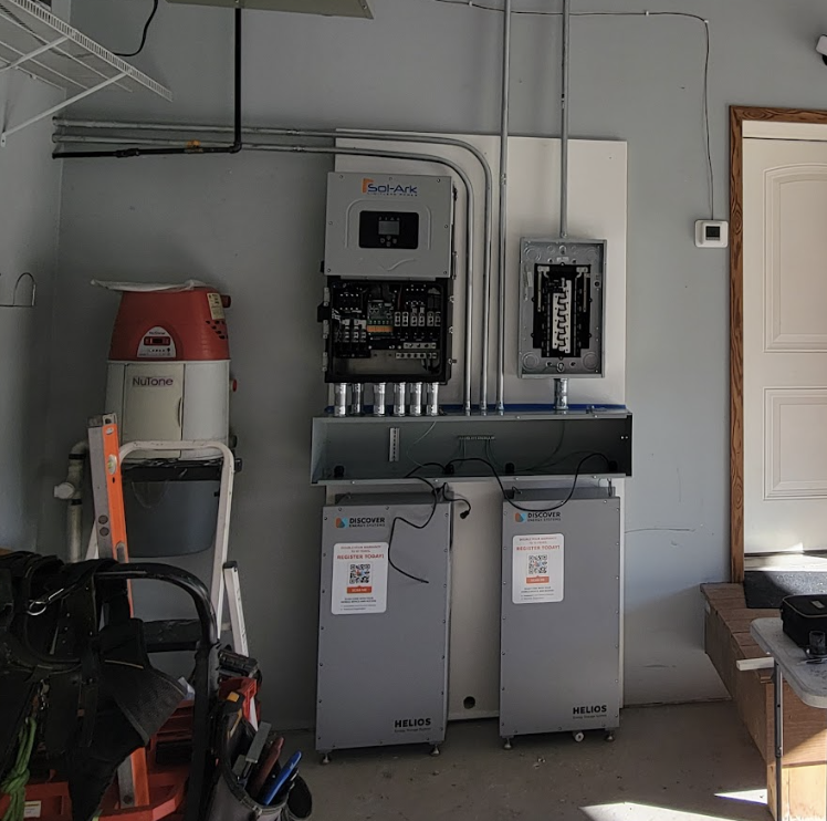 Indoor utility room with a solar inverter on the wall and two gray Helios battery cabinets below; exposed wiring and a ladder nearby, tools on a cart in the foreground.