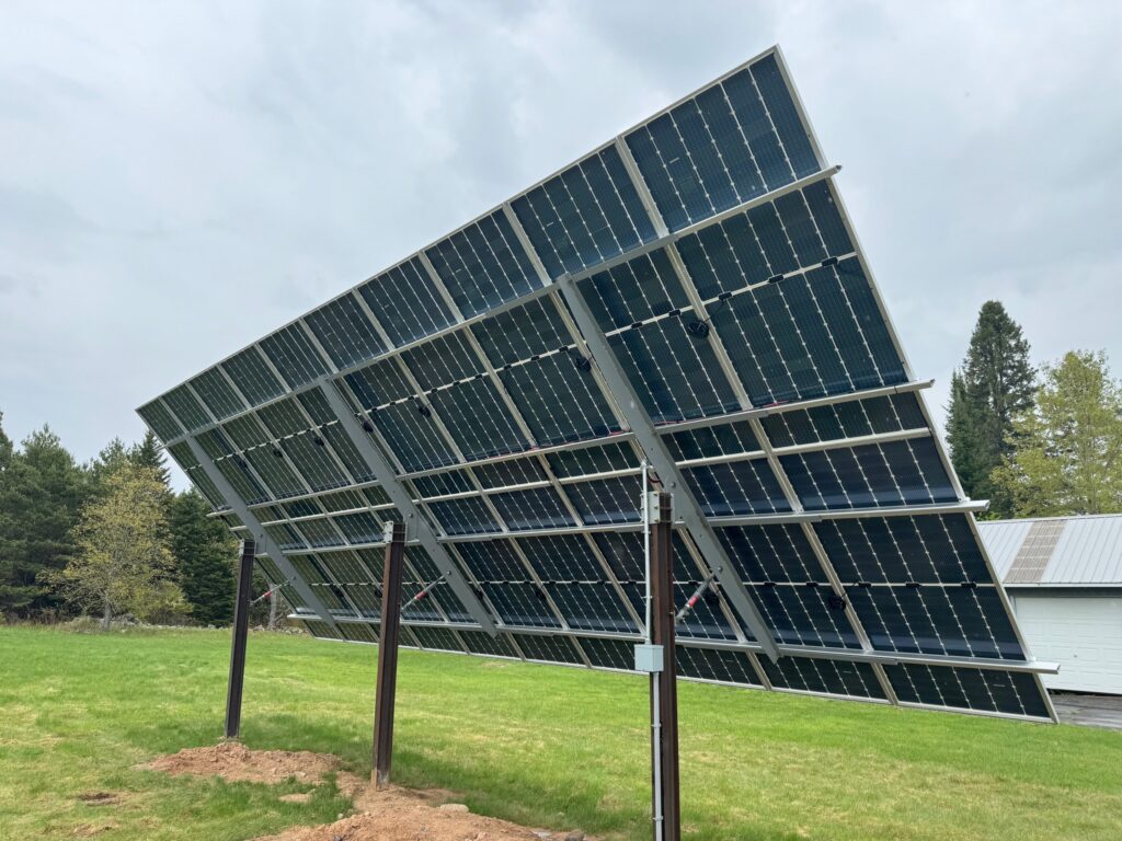 Tilted solar panel array mounted on metal frames in a grassy field with trees and a cloudy sky behind it by a building edge.
