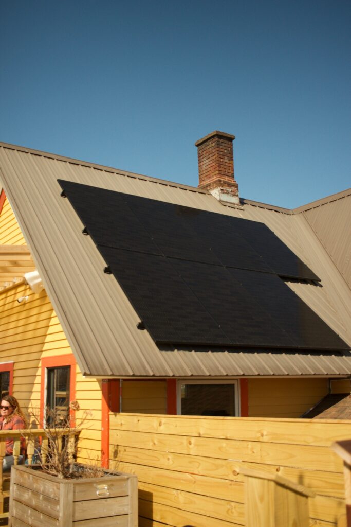 House roof with dark solar panels installed on a light metal roof under a clear blue sky; brick chimney rises above.
