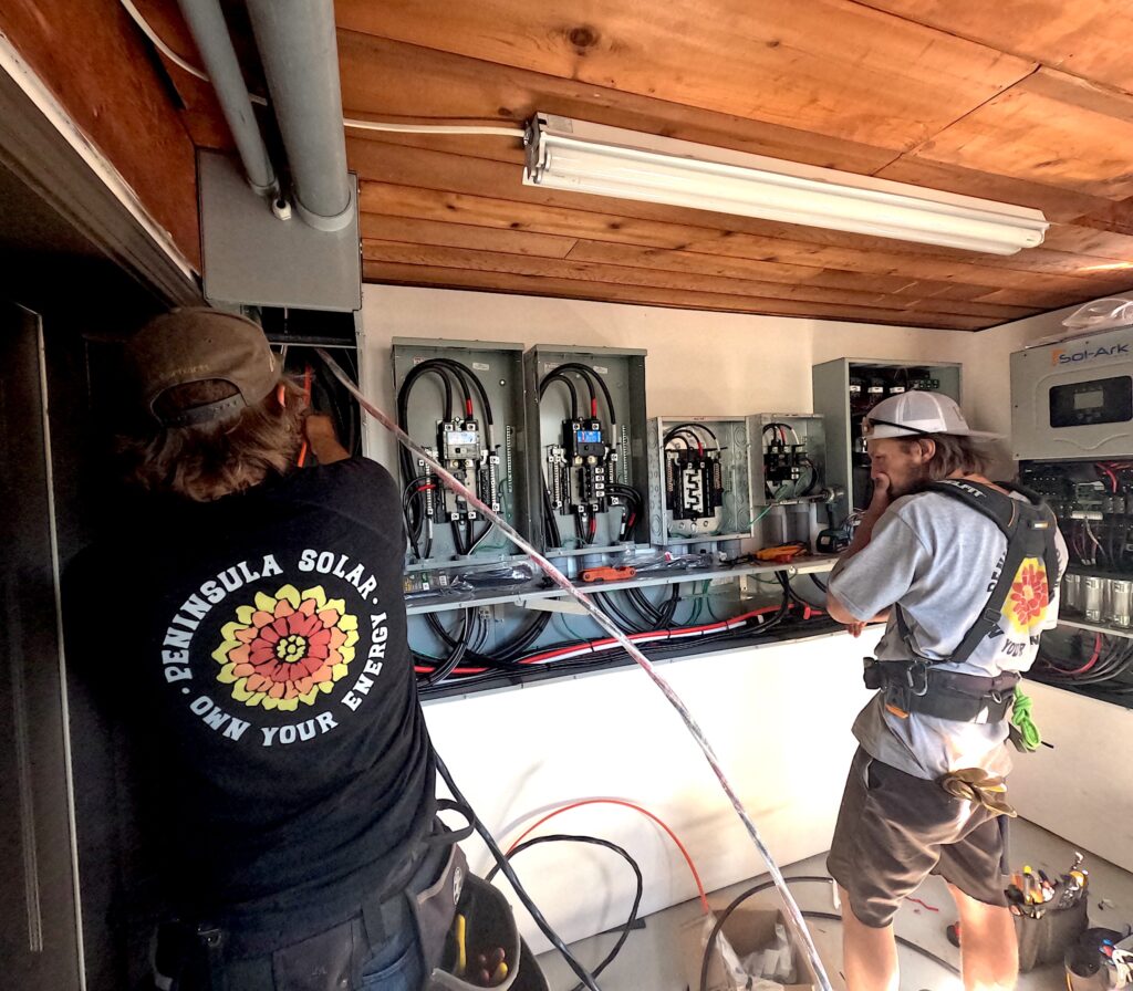 DCIM100GOPROG0040147JPG – Peninsula Solar Two technicians in work shirts and hats wiring open electrical panels in a utility room with an exposed wooden ceiling conduit pipes nearby.