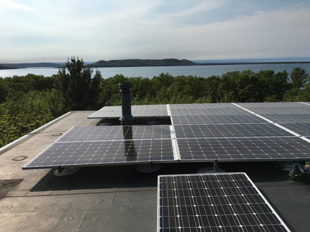 Peninsula-solar-solar-panels-producing-energy-on-sunny-summer-day-northern-michigan – Peninsula Solar Solar panels on a flat roof overlook a lake with forested hills in the distance.
