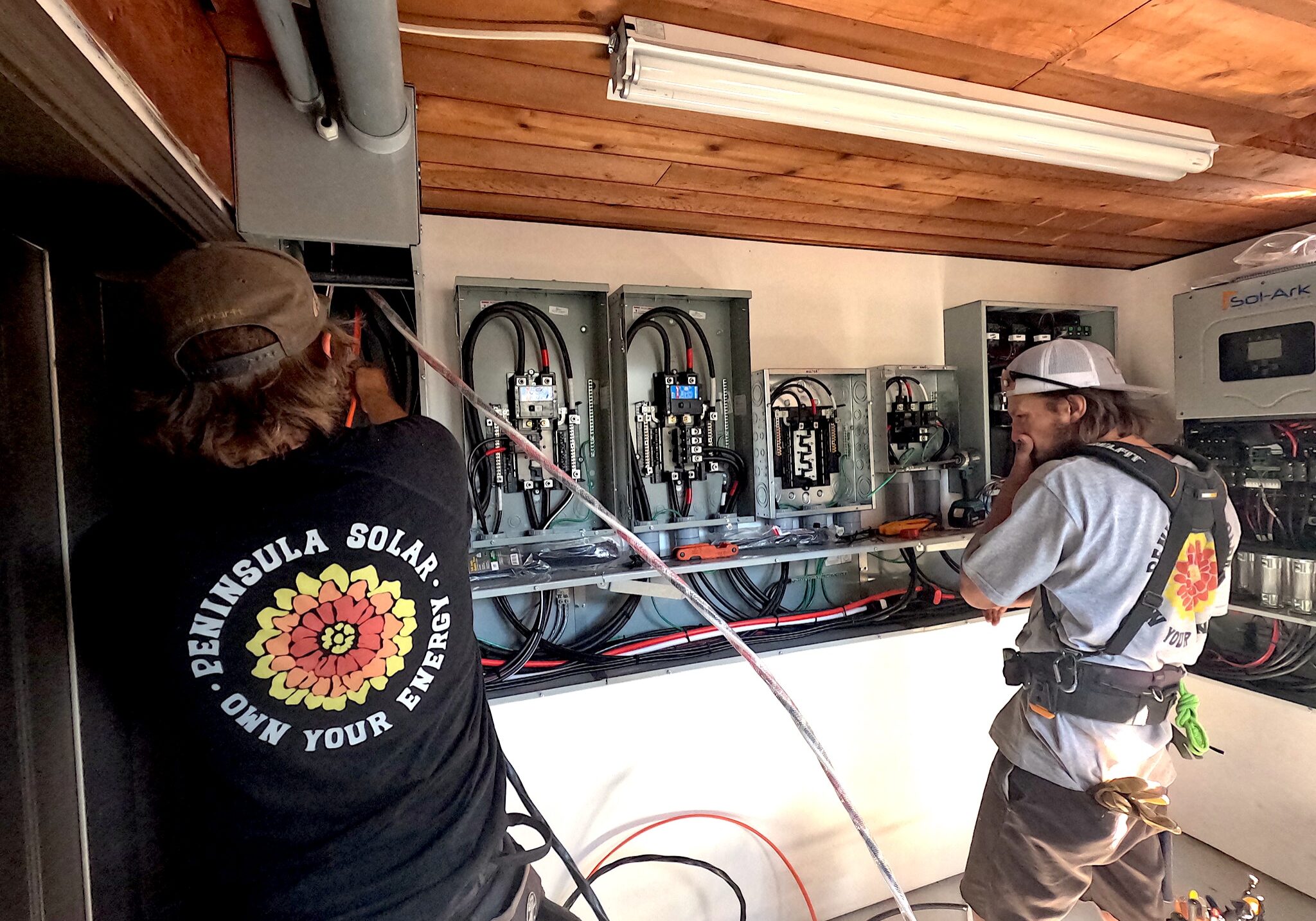 Two technicians in work shirts and hats wiring open electrical panels in a utility room with an exposed wooden ceiling conduit pipes nearby.